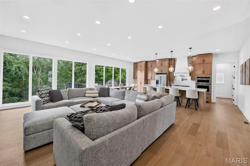 Open-concept living space featuring wood-finish flooring, recessed lighting, expansive windows, and a kitchen with warm-toned cabinetry and a center island with seating — 3 Millers Court, O'Fallon, MO 63366
