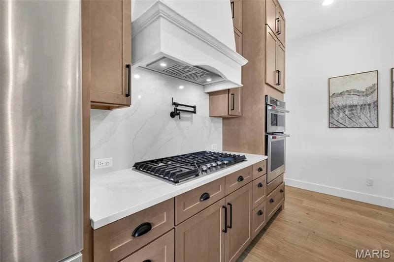 Contemporary kitchen featuring custom wood-finish cabinetry with integrated hardware, white quartz countertops, professional-grade gas cooktop, stainless steel refrigerator, built-in range hood with white surround, and hardwood flooring throughout — 3 Millers Court, O'Fallon, MO 63366