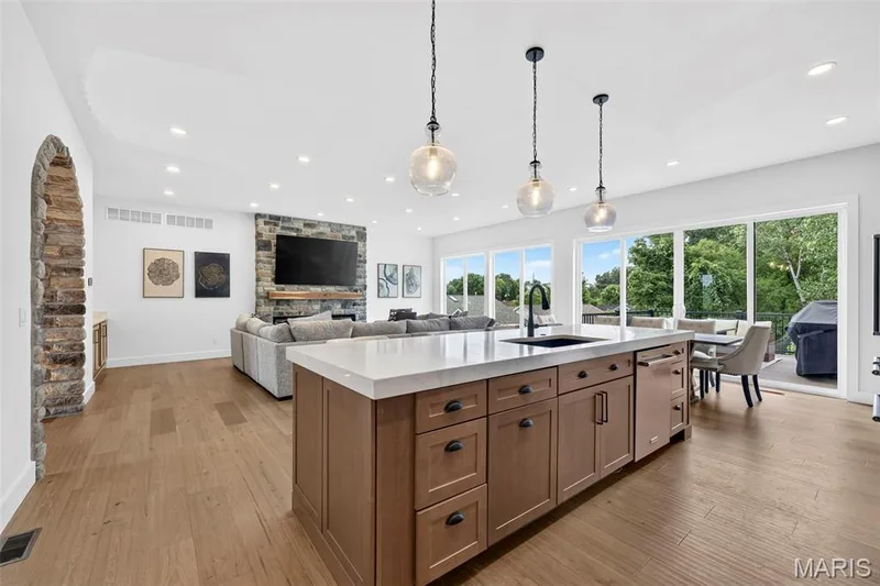 Open-concept kitchen and living area featuring a wood-finish island with multiple drawers and cabinet storage, quartz countertops, pendant lighting, wood-finish flooring throughout, and expansive glass doors and windows framing landscaped grounds and mature trees — 3 Millers Court, O'Fallon, MO 63366