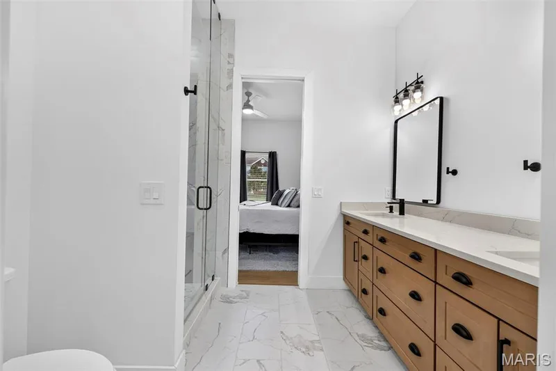 Primary bathroom featuring a wood-finish dual-sink vanity with black hardware, white quartz countertop, framed mirror with integrated lighting, and tile flooring — 3 Millers Court, O'Fallon, MO 63366