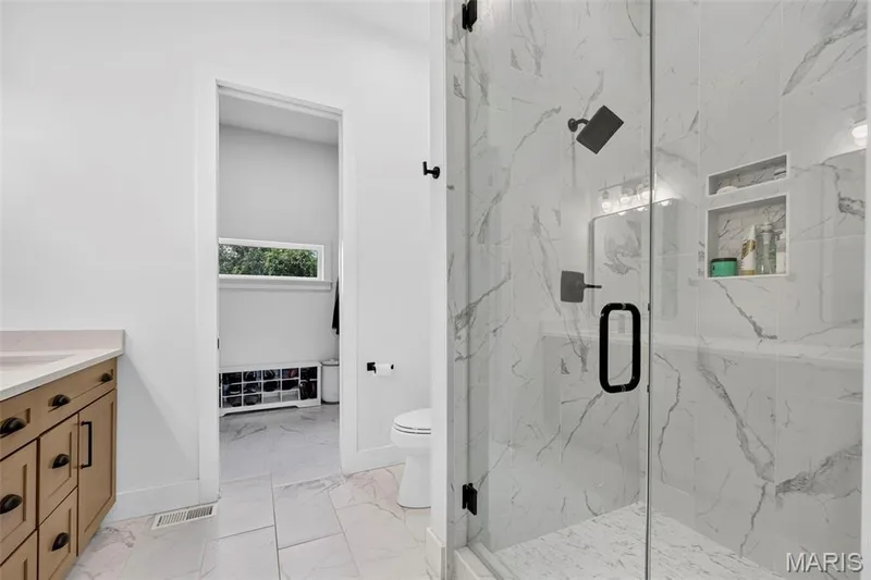 Spa-inspired primary bathroom featuring a frameless glass-enclosed shower with marble-look tile surround, polished marble-look flooring, and a wood-finish vanity with integrated sink and hardware — 3 Millers Court, O'Fallon, MO 63366