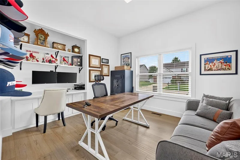 Bright home office featuring wood-finish flooring, a built-in desk with white cabinetry and shelving, large windows with plantation shutters providing natural light, and a comfortable seating area with upholstered furnishings positioned for versatile use — 3 Millers Court, O'Fallon, MO 63366