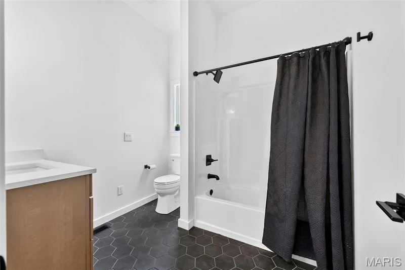 Contemporary bathroom featuring a wood-finish vanity with integrated sink, white subway tile surround, hexagonal tile flooring in charcoal, and a black metal shower rod with dark curtain — 3 Millers Court, O'Fallon, MO 63366