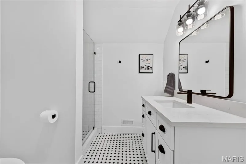 Modern primary bathroom featuring a dual-sink vanity with white cabinetry and ample countertop space, large framed mirror with integrated lighting, black metal fixtures and hardware, black-and-white geometric tile flooring, and a separate walk-in shower with glass enclosure and subway tile walls — 3 Millers Court, O'Fallon, MO 63366