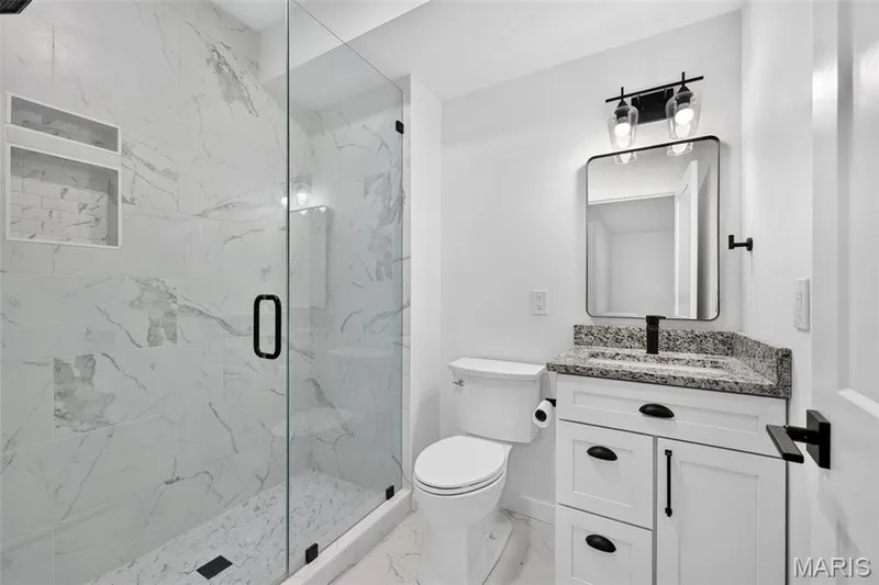 Spa-inspired bathroom featuring a walk-in shower with marble-look tiled walls and frameless glass enclosure, white vanity with dual drawers and brushed nickel hardware, granite-look countertop with integrated sink, and polished chrome fixtures — 3 Millers Court, O'Fallon, MO 63366