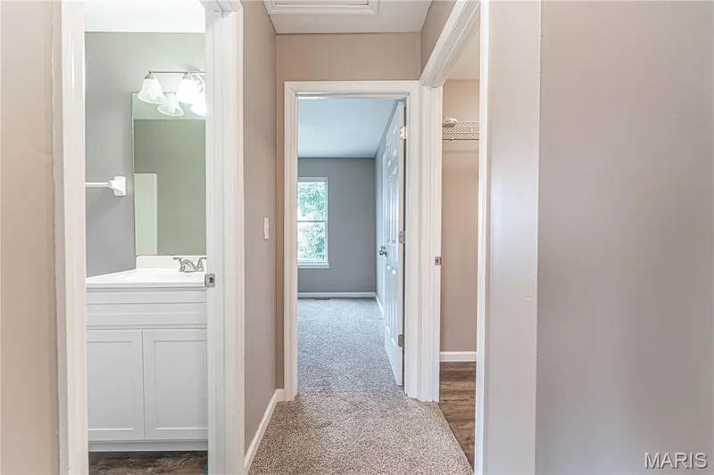 Carpeted hallway with painted walls, leading to a room with a window and a bathroom with a white vanity and mirror — 1721 Summergate Court, St Charles, MO 63303