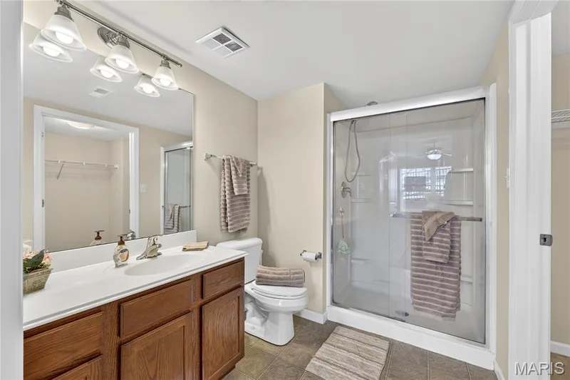 Full bathroom featuring a wood-finish vanity with a single sink, a large wall-mounted mirror, a glass-enclosed shower, and neutral-toned wall paint — 313 Bramblett Hills, O'Fallon, MO 63366
