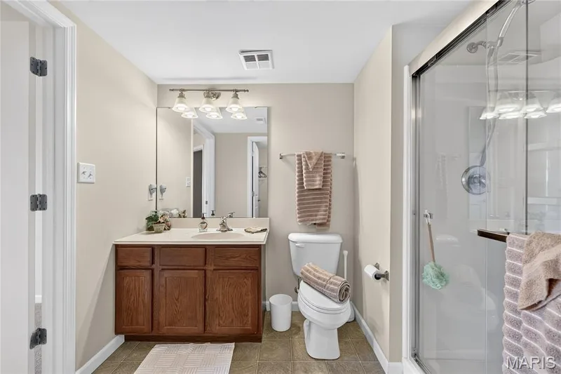 Bathroom featuring a glass-enclosed shower, wood-finish vanity with countertop, wall-mounted mirror, and overhead vanity lighting — 313 Bramblett Hills, O'Fallon, MO 63366