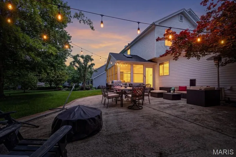 Spacious concrete patio featuring string lighting, an enclosed sunroom, a vibrant red tree, and a large grassy area — 17821 Wilderness Cliff Court, Wildwood, MO 63005
