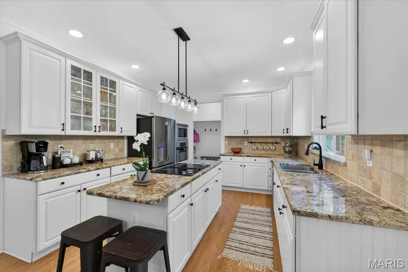 Bright kitchen featuring a central island with a built-in cooktop, light wood-finish flooring, white cabinetry with black hardware, granite countertops, and a tiled backsplash — 17821 Wilderness Cliff Court, Wildwood, MO 63005