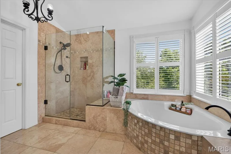 Spacious bathroom featuring a built-in soaking tub with mosaic tile surround, an enclosed glass shower with tiled walls and a built-in niche, and large windows with plantation shutters — 17821 Wilderness Cliff Court, Wildwood, MO 63005