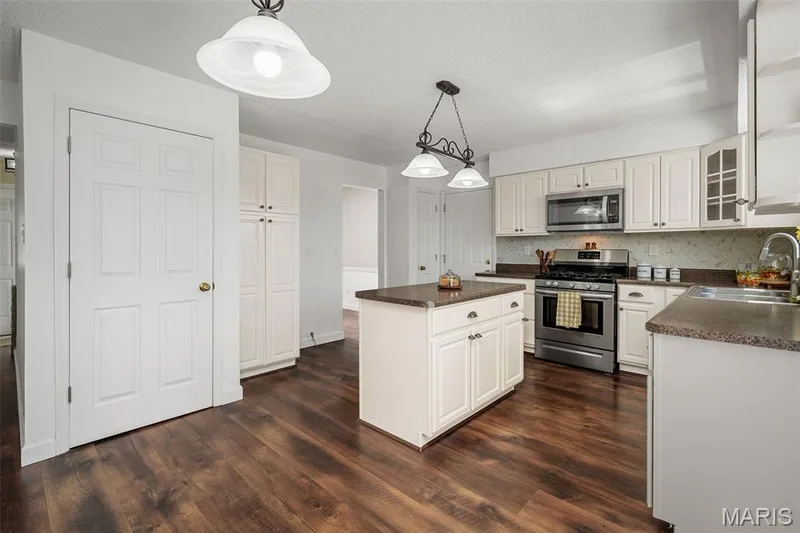 Spacious kitchen featuring cream cabinetry with brushed nickel hardware, stainless steel gas range and microwave, wood-finish flooring throughout, a centered island with prep space, and pendant lighting suspended from the ceiling — 120 Bromley Court, O'Fallon, MO 63368