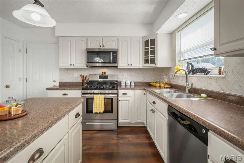 Kitchen featuring stainless steel appliances, white cabinetry, dark wood-finish flooring, a patterned tile backsplash, and a double basin sink — 120 Bromley Court, O'Fallon, MO 63368