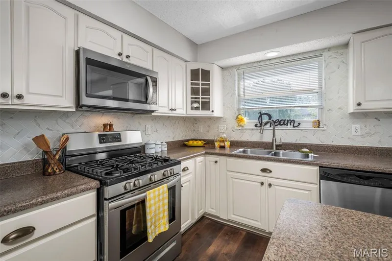 White cabinetry with bronze hardware, stainless steel appliances, herringbone tile backsplash, double basin sink, and wood-finish flooring — 120 Bromley Court, O'Fallon, MO 63368
