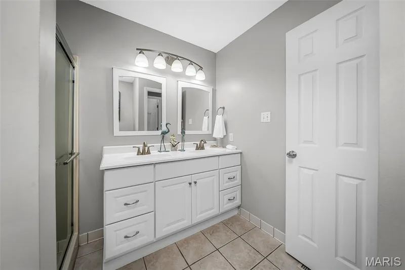 Dual vanity with white cabinetry, sleek countertops, multiple mirrors, and a glass-enclosed shower — 120 Bromley Court, O'Fallon, MO 63368