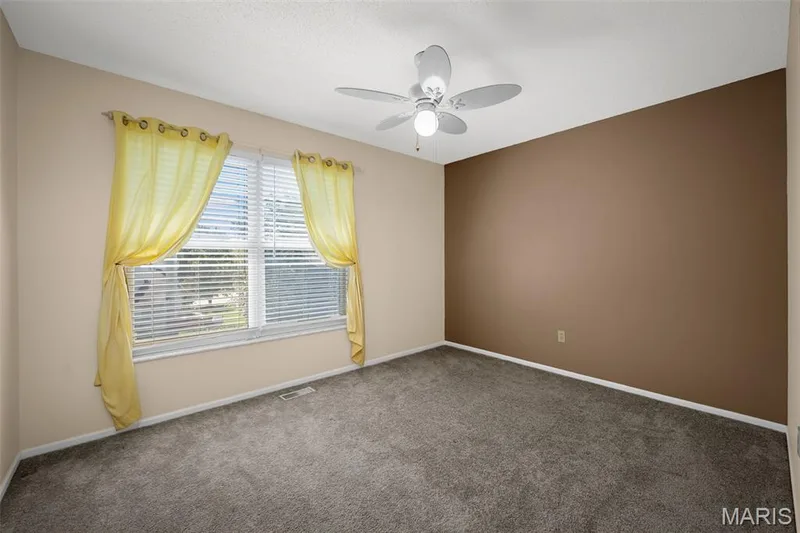Spacious bedroom offering neutral wall tones, plush carpeting, ceiling fan with integrated lighting, and large windows dressed in yellow grommet curtains that flood the room with natural light — 120 Bromley Court, O'Fallon, MO 63368