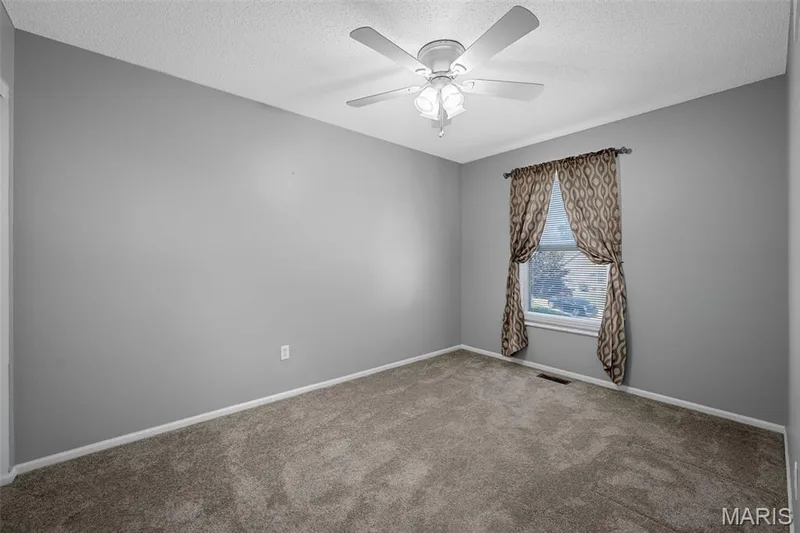 Neutral tone wall paint, textured ceiling, ceiling fan with light fixture, single window with patterned drapes, and carpet flooring — 120 Bromley Court, O'Fallon, MO 63368