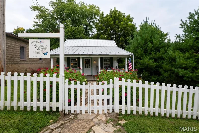 White picket fence with a gate, stone walkway, and established flowers/ landscaping — 215 Jackson Street, Augusta, MO 63332