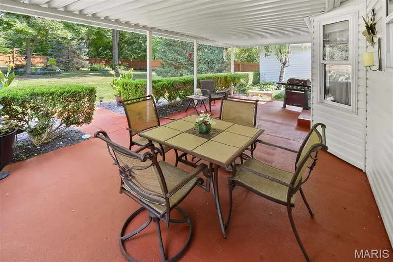 Covered outdoor patio with a red-toned concrete surface, surrounded by manicured shrubs — 38 Fawn Valley Circle, St Peters, MO 63376