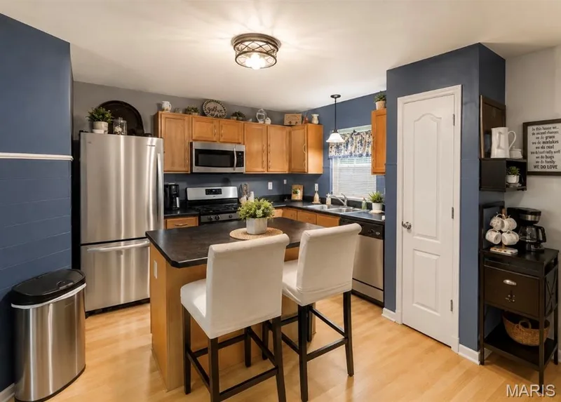 Modern kitchen featuring warm wood cabinetry, stainless steel appliances including a microwave and refrigerator, black cooktop, and wood-finish flooring — 3231 Cog Wheel Station, St Charles, MO 63301