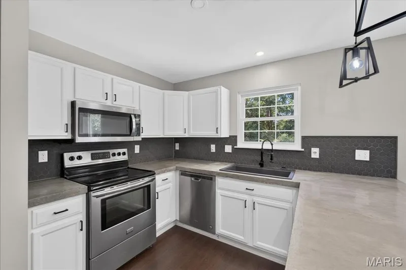 White cabinetry with black hardware, stainless steel appliances, dark hexagonal tile backsplash, concrete-finish countertops, and wood-finish flooring — 53 Lions Head Court, O'Fallon, MO 63368