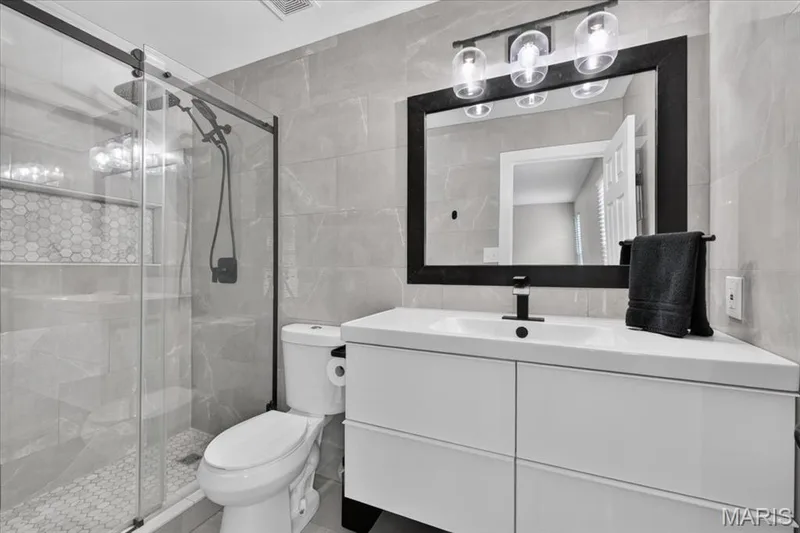 Glass-enclosed shower with a hex-tiled floor, a built-in shelf, and a black-framed mirror over a white vanity with an integrated sink — 53 Lions Head Court, O'Fallon, MO 63368