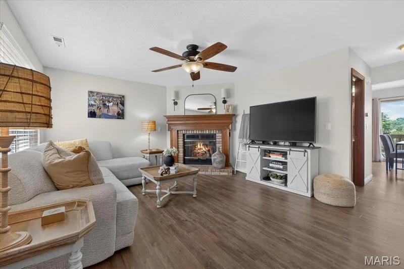 Wood-finish flooring throughout, a corner fireplace with a wood mantle and brick surround, and a ceiling fan with integrated lighting — 1383 Forest Creek Drive #5, St Charles, MO 63303