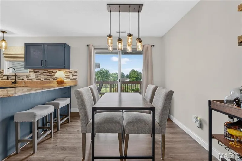 Open-concept dining and kitchen area featuring a wood-finish plank floor, pendant lighting suspended from a metal fixture, and expansive glass doors opening to an outdoor terrace with tree-lined landscape beyond — 1383 Forest Creek Drive #5, St Charles, MO 63303