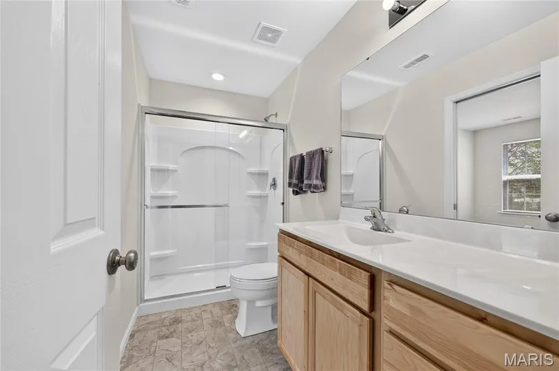 Shower with clear glass enclosure and integrated shelving, vanity with light wood finish cabinetry, white countertop with integrated sink, and large wall-mounted mirror — 352 Fall Harvest Parkway, Wentzville, MO 63385