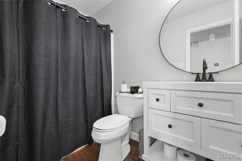 White vanity with black hardware, circular mirror, black faucet, and wood-finish flooring — 1094 Treeshade Drive, St Peters, MO 63376