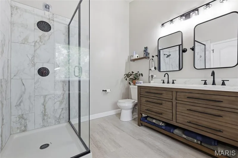 Glass-enclosed shower with tile surround, wood-finish double vanity with dual sinks, and two framed mirrors — 1094 Treeshade Drive, St Peters, MO 63376