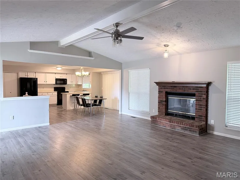 Vaulted ceiling with exposed beams, wood-finish flooring, brick fireplace with mantel, white cabinetry, and black appliances — 21 White Chapel, O'Fallon, MO 63368