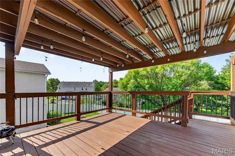 Expansive covered deck featuring wood-finish flooring, wood railings with black balusters, and a corrugated metal ceiling — 934 Annabrook Park Drive, O'Fallon, MO 63366