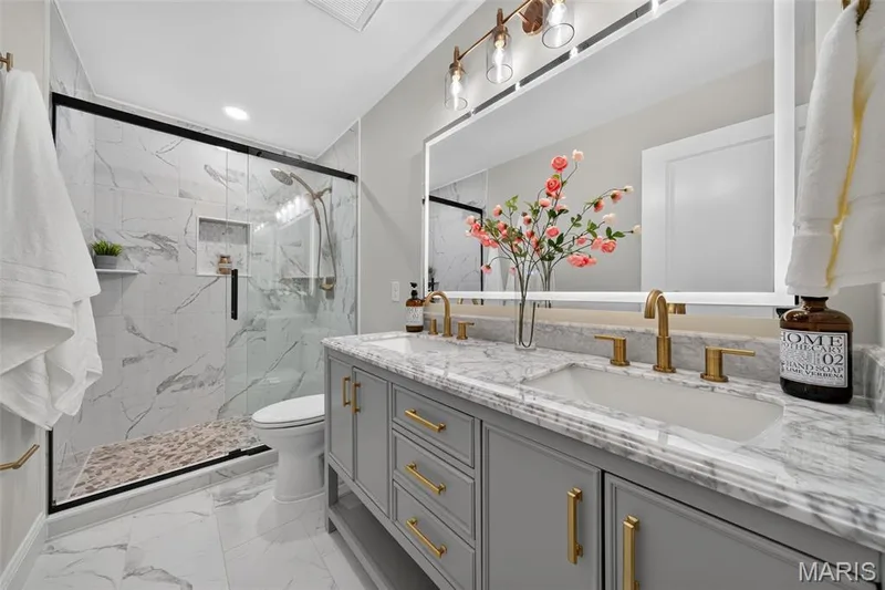 The primary bath features dual vanities, a tiled shower, layered lighting, and champagne-bronze finishes. — 11075 Village Drive, Foristell, MO 63348