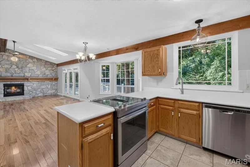 Vaulted ceiling with exposed wood beams, stone-clad fireplace with wood mantle, wood-finish flooring, white kitchen countertops, and stainless steel kitchen appliances — 20 Ardennais Road, St Peters, MO 63376