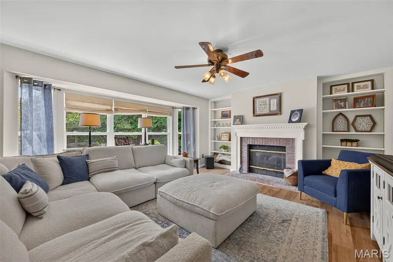 Bay window with natural light, brick fireplace with mantel, built-in shelving, wood-finish flooring, and a ceiling fan with integrated lighting — 10 Docs Ct., St Charles, MO 63303