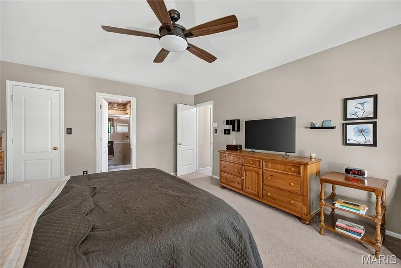 Spacious primary bedroom featuring neutral walls, ceiling fan with light fixture, wood-finish dresser with ample storage, carpeted flooring, and multiple doorways providing access to ensuite and walk-in closet areas — 10 Docs Ct., St Charles, MO 63303