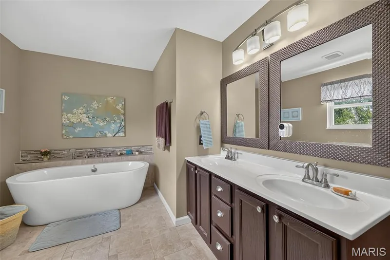 Freestanding soaking tub, double vanity with integrated sinks, framed mirrors, wall-mounted light fixture, and tiled flooring — 10 Docs Ct., St Charles, MO 63303