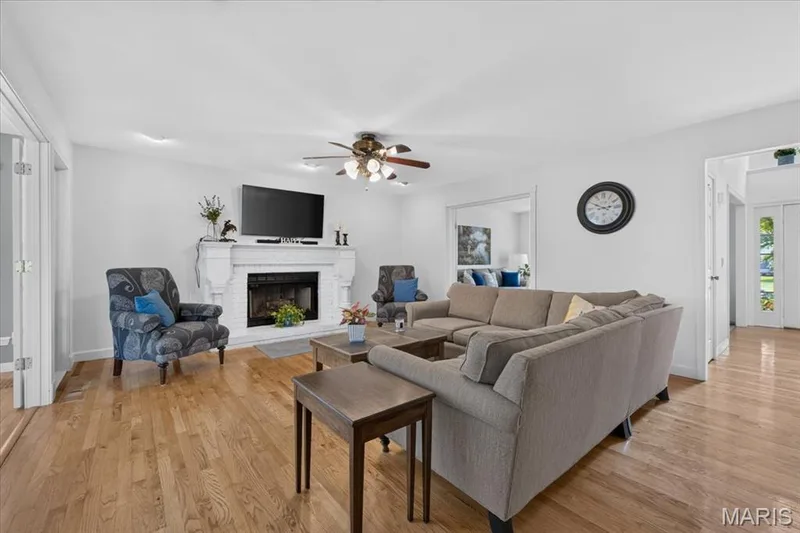 Expansive living area featuring wood-finish flooring, a white brick fireplace with mantel, and a ceiling fan — 5856 Canterfield Court, Weldon Spring, MO 63304