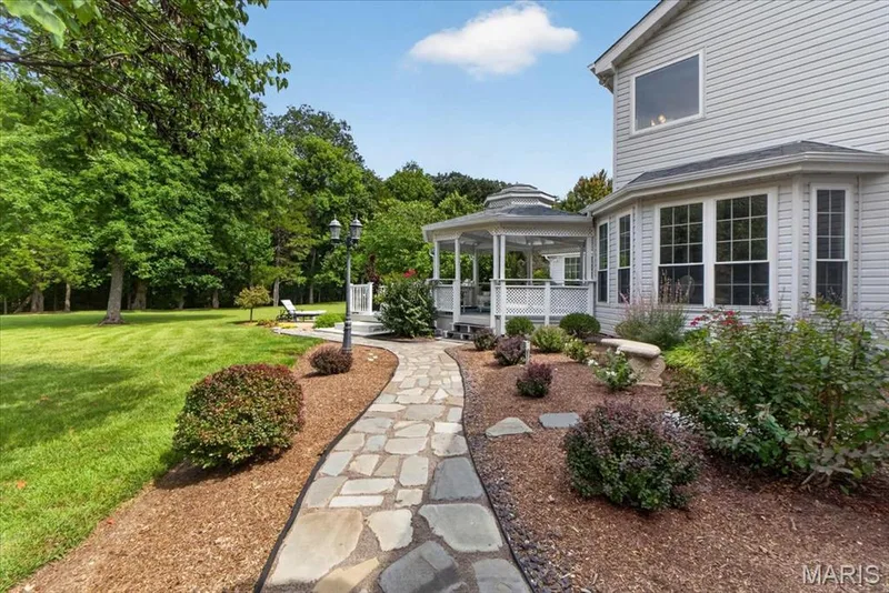 Stone pathway leading to a hexagonal gazebo, surrounded by mature trees, manicured lawn, and landscaped garden beds — 5856 Canterfield Court, Weldon Spring, MO 63304