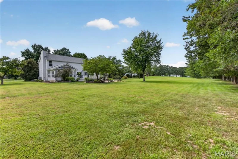 Expansive green lawn with mature trees and a two-story residence featuring a porch and chimney — 5856 Canterfield Court, Weldon Spring, MO 63304