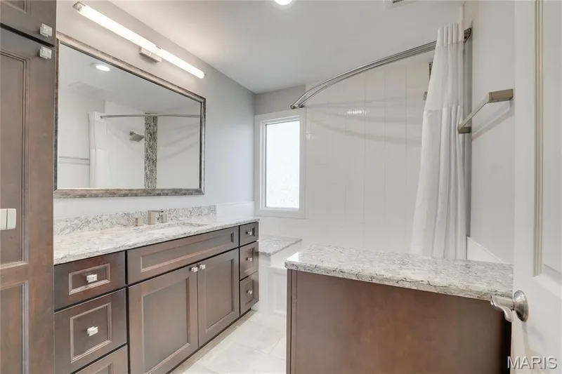 A wood-finish vanity with multiple drawers and a marble-look countertop, large framed mirror with integrated lighting, and a walk-in shower with glass enclosure and marble-look surround — 2508 Peppermill Lake Court, Chesterfield, MO 63005