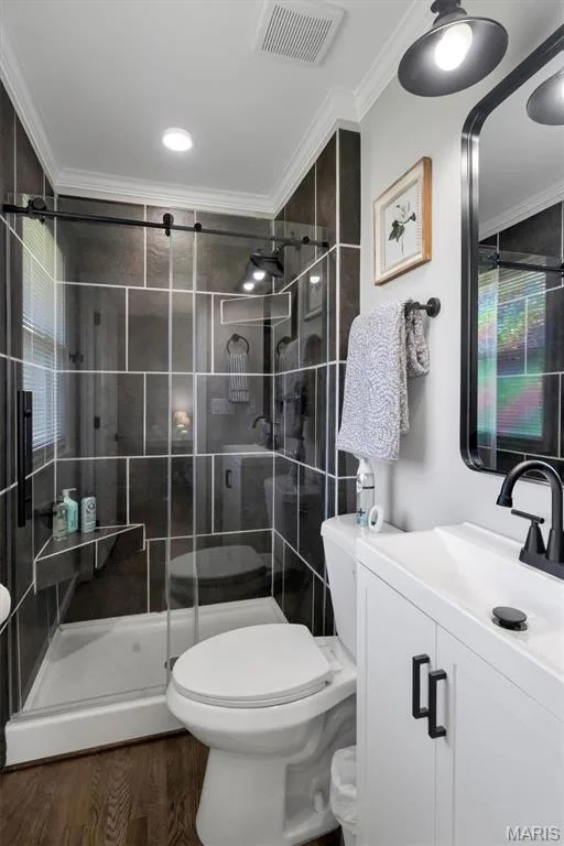 Walk-in shower with dark tile surround and glass enclosure, white vanity with black hardware, wood-finish flooring, and crown molding — 7 Saint Mark Drive, St Peters, MO 63376