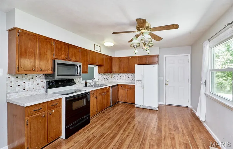 Kitchen featuring wood-finish flooring, wood cabinetry, a mosaic tile backsplash, stainless steel microwave, and white appliances — 318 Capri Drive, O'Fallon, MO 63366