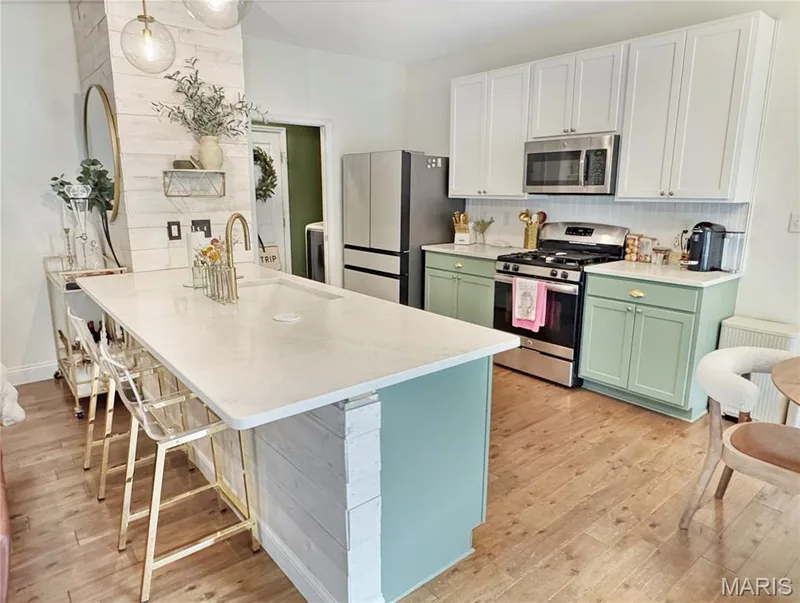 Kitchen featuring wood-finish flooring, two-tone cabinetry, stainless steel appliances, a white tile backsplash, and a large island with an integrated sink and gold-tone faucet — 4024 Glengarry Drive, Wentzville, MO 63385