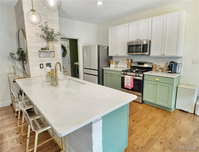Kitchen featuring wood-finish flooring, a central island with a built-in sink, white stone-finish countertops, two-tone cabinetry, and stainless steel appliances — 4024 Glengarry Drive, Wentzville, MO 63385