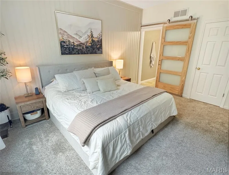 Soft gray carpet flooring, beadboard wall paneling, tufted fabric headboard, wood-frame barn door with frosted glass panels, and a standard paneled interior door — 4024 Glengarry Drive, Wentzville, MO 63385