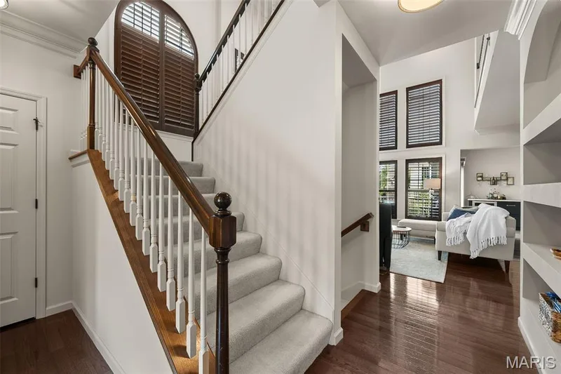 Hardwood flooring, carpeted staircase with wood handrails and white balusters, arched window with plantation shutters, multiple large windows with plantation shutters, and built-in shelving — 2700 Grover Crossing Drive, Wildwood, MO 63040