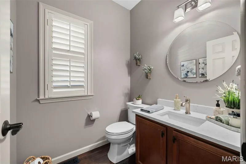 Window with white plantation shutters, vanity with white countertop, integrated sink, and wood-finish cabinetry, oval frameless mirror, and a two-bulb vanity light fixture — 2700 Grover Crossing Drive, Wildwood, MO 63040