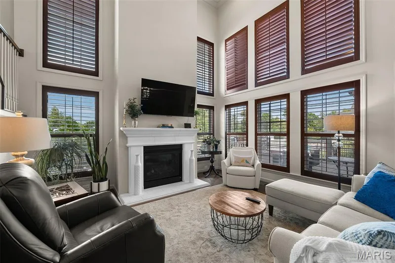 Living area featuring a floor-to-ceiling fireplace with a white mantel, high ceilings, multiple windows with dark wood frames, and a staircase with white balusters — 2700 Grover Crossing Drive, Wildwood, MO 63040